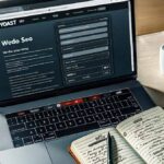Foto de um laptop aberto em uma página de edição de site, com a interface do plugin YoastSEO exibida proeminentemente na tela. Ao lado do laptop, um bloco de notas com dicas de SEO escritas à mão e uma xícara de café.