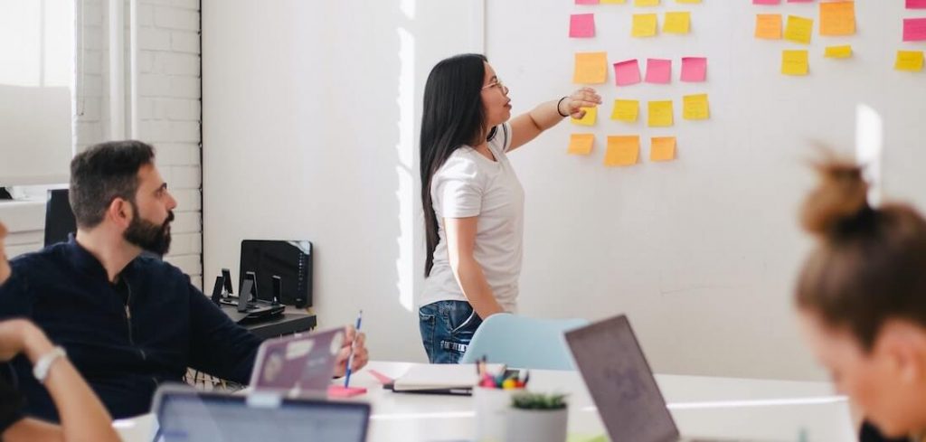 mulher aponta para post its em um quadro branco enquanto grupo reunido em volta de mesa dá suporte